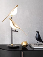 Bellvian golden bird LED table lamp on desk beside plant, warm lighting for home office or reading corner.