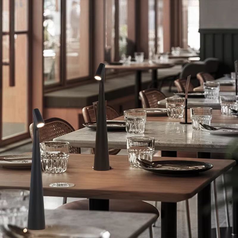 Modern black LED table lamps placed on elegant wooden dining tables inside a stylish restaurant. The lamps emit a soft warm light that enhances the cozy dining atmosphere, highlighting minimalist decor with glassware and neutral tones. Perfect ambient lighting for luxury cafes and restaurants.