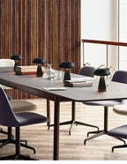 Set of matte black mushroom LED table lamps placed on a modern conference table surrounded by grey chairs. Cordless, rechargeable lamps providing ambient and task lighting for offices, cafés, or meeting spaces.