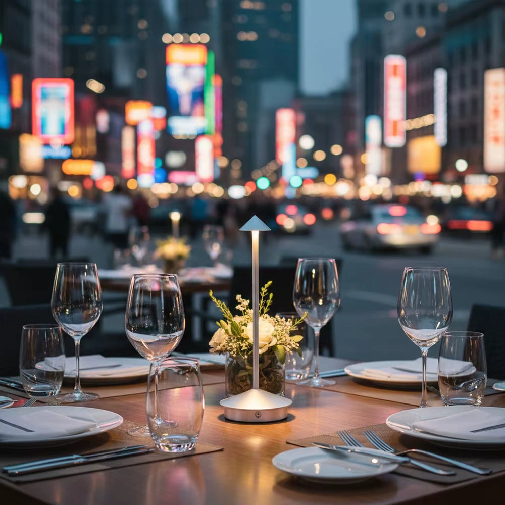 Cordless LED table lamp glowing softly on a restaurant table with glassware and flowers, surrounded by city lights and blurred night traffic in the background — perfect modern ambient lighting for outdoor dining.