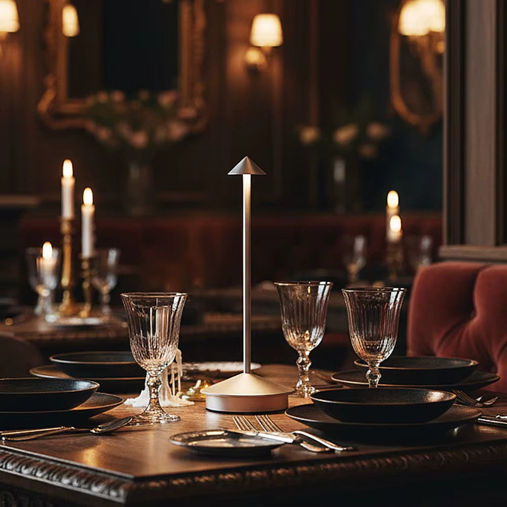 Minimalist cordless LED table lamp placed on an elegant dining table with crystal glasses, dark plates, and candles in a luxury restaurant setting, creating warm ambient lighting and refined atmosphere.