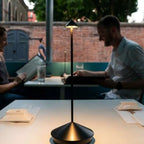Cordless LED table lamp glowing on a restaurant table while two people dine outdoors, creating a warm and intimate evening ambiance.