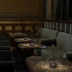 Elegant cordless LED lamps arranged on marble tables in a luxury restaurant interior with dark green seating and warm ambient lighting.