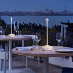Cordless LED lamps illuminating outdoor restaurant tables with glassware and plates, overlooking a city waterfront at night.