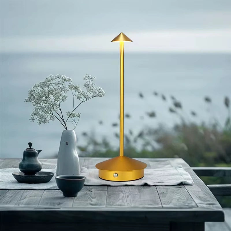 Modern golden cordless LED lamp on a wooden table with tea set and flower vase, overlooking a peaceful seaside background.
