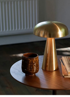 Gold metal mushroom LED table lamp placed on a round wooden table beside a ceramic mug. Warm ambient lighting creates a cosy and elegant home atmosphere, ideal for bedrooms, reading corners, and minimalist interiors.