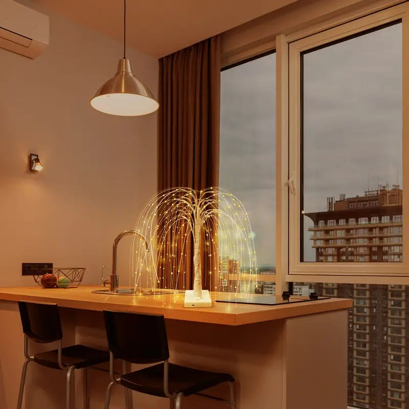 LED willow tree lamp with warm yellow lights glowing on a kitchen counter beside large windows overlooking city buildings. The soft ambient light enhances modern interior design, creating a cozy and elegant evening atmosphere.