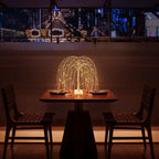Romantic LED willow tree lamp glowing on a wooden dining table set for two in a dimly lit restaurant. The cascading golden fairy lights create a warm, intimate atmosphere ideal for dinners, cafes, or cozy home lighting decor. Bellvian