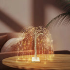 Elegant LED willow tree lamp placed on a round wooden table in a cozy living room. The golden fairy lights cascade from flexible branches, creating a warm ambient glow. Perfect for home decoration, relaxation lighting, or modern minimalist interiors.