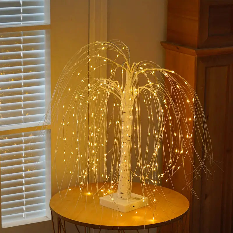 LED willow tree lamp positioned near a window, glowing softly in a warm-toned room. The cascading lights and white trunk add an elegant, nature-inspired touch to modern home interiors and evening lighting decor. Bellvian