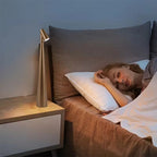 Modern minimalist bedside LED lamp emitting warm light beside a sleeping woman. The elegant metallic gold lamp stands on a wooden nightstand next to a soft brown bed, creating a cozy and relaxing bedroom atmosphere.